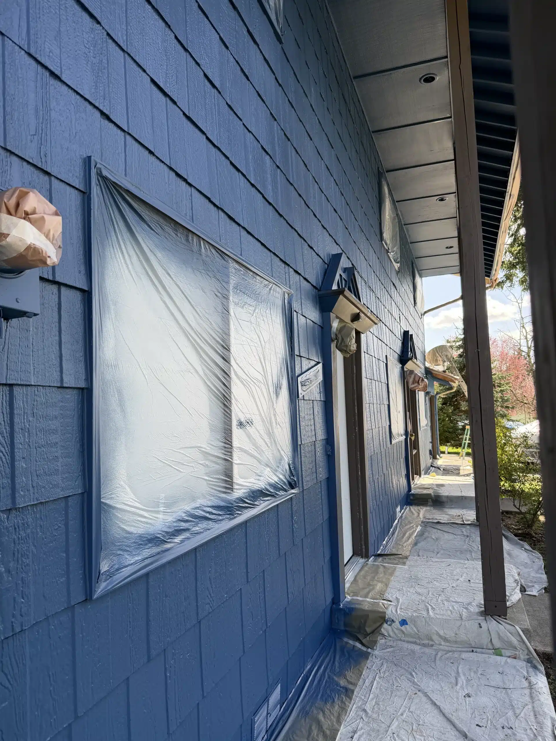 Professional Exterior House Painting Services Exterior painting in progress on a blue house with windows protected by plastic sheeting and drop cloths.