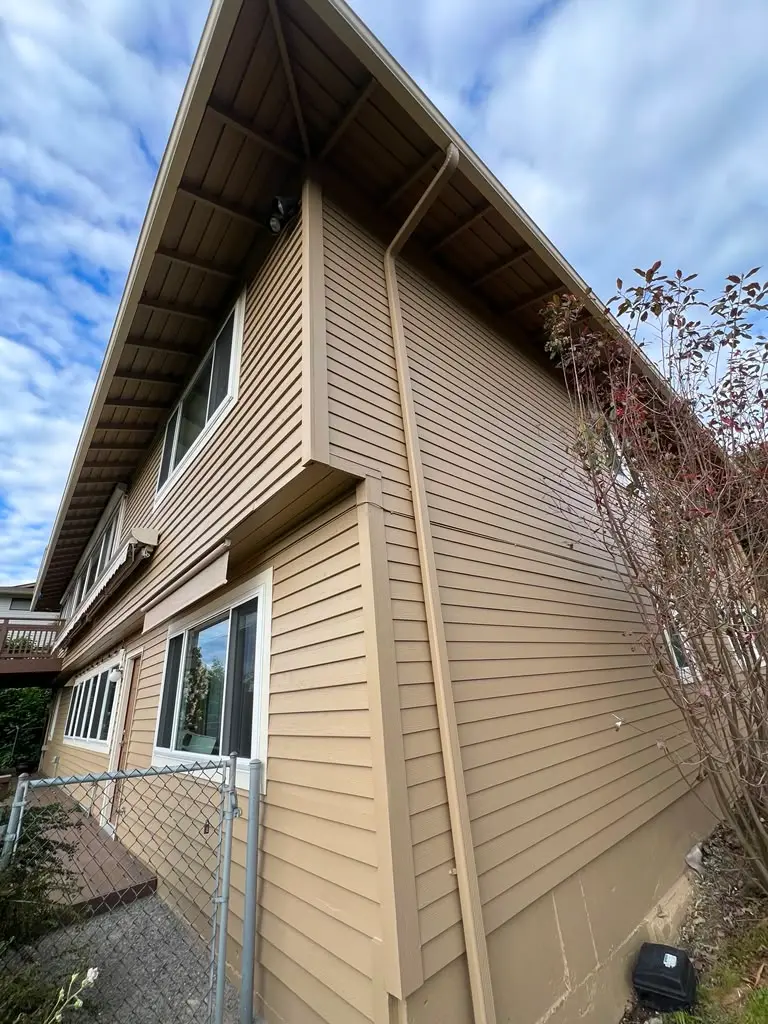 House Exterior Painting Services for Siding and Trim Side view of a residential house with light brown siding and white trim under an overcast sky.