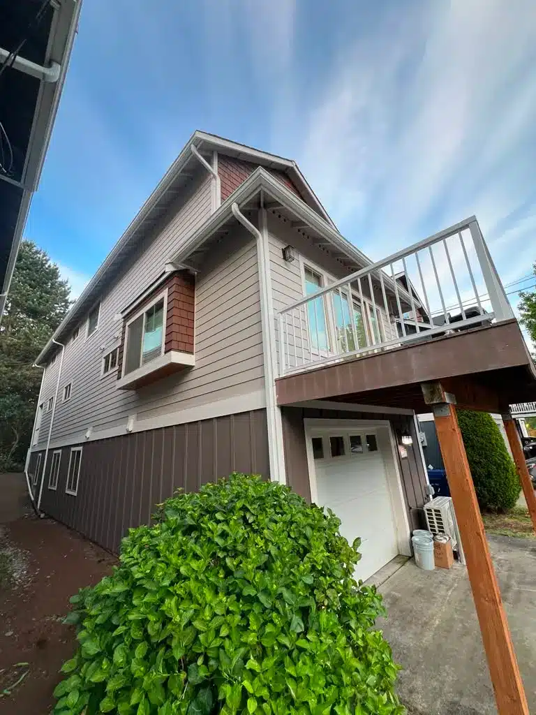 Exterior House Painting with Siding Trim and Garage Doors Side view of a residential house featuring gray siding, brown shingles, a white garage door, and an upper deck.